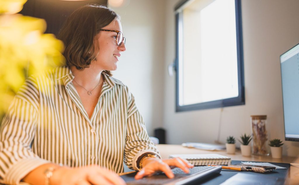 Smiling woman working on a desktop computer in a bright office, wearing a striped shirt and glasses, with plants and a notebook on her desk