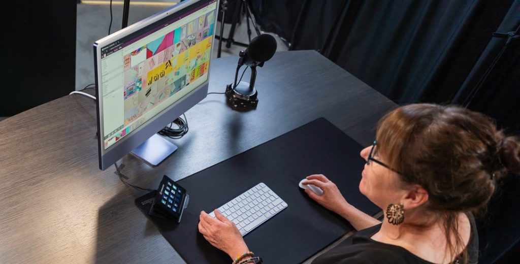 Woman using a desktop computer with a large screen displaying a grid of coloured visuals, in a workspace equipped with a microphone and a Stream Deck