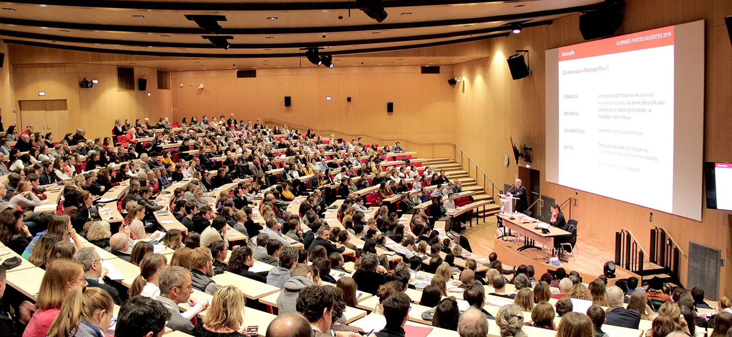 Amphitheatre where hundreds of students listen to two professors speaking on their platform.
