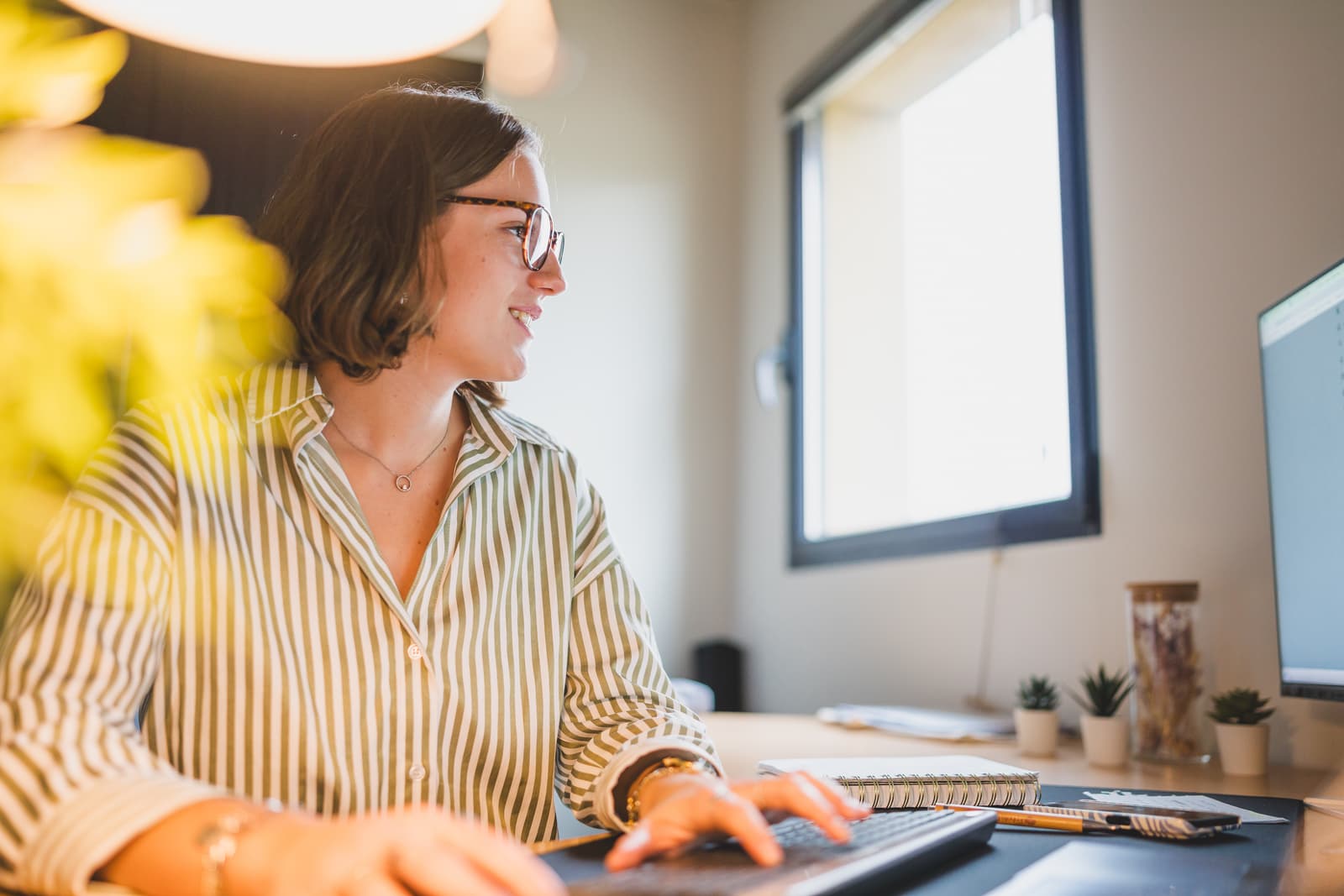 Woman wearing glasses in front of her computer, looking at her screen and typing on the keyboard.
