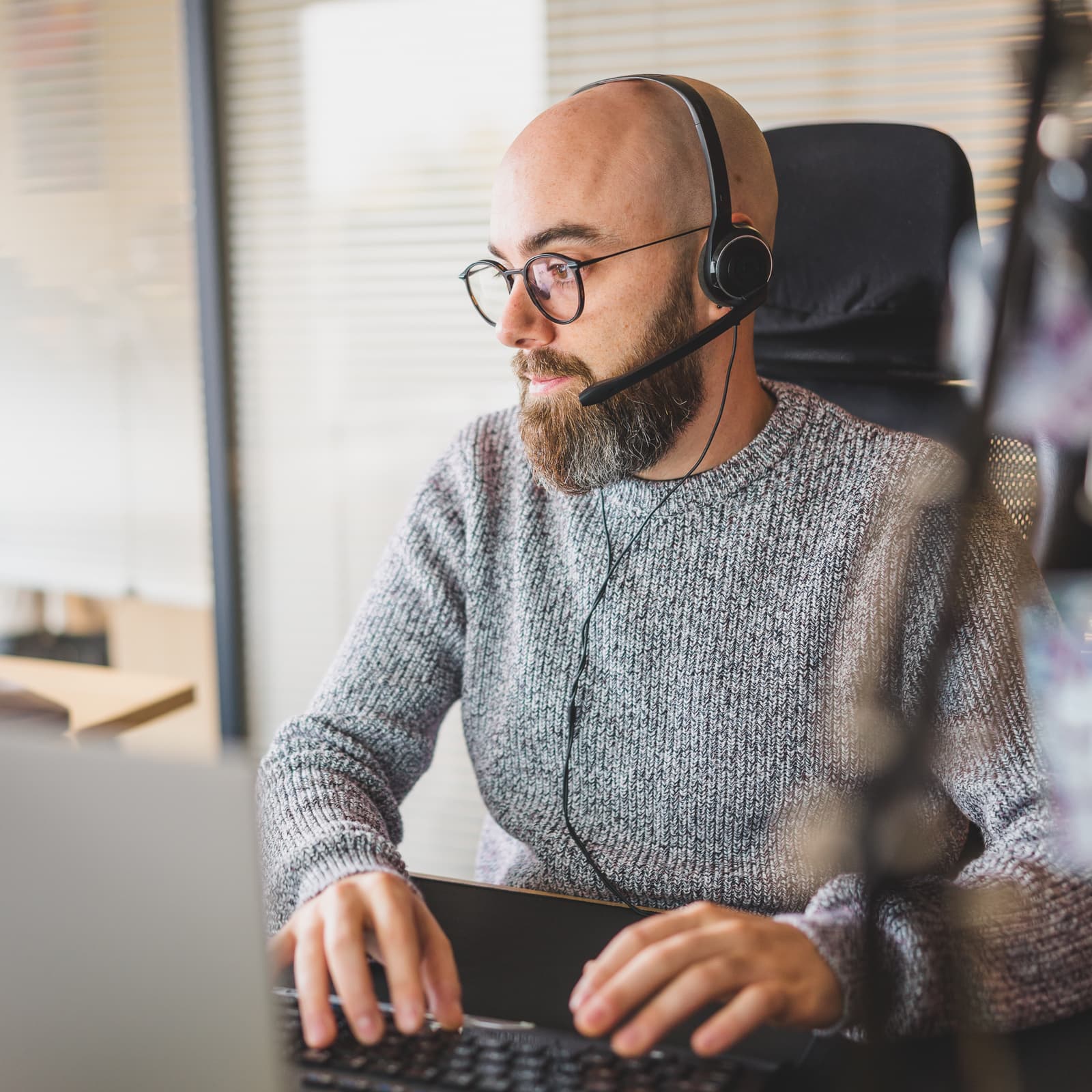 Man with glasses wearing headphones looking at his computer and typing on his keyboard.