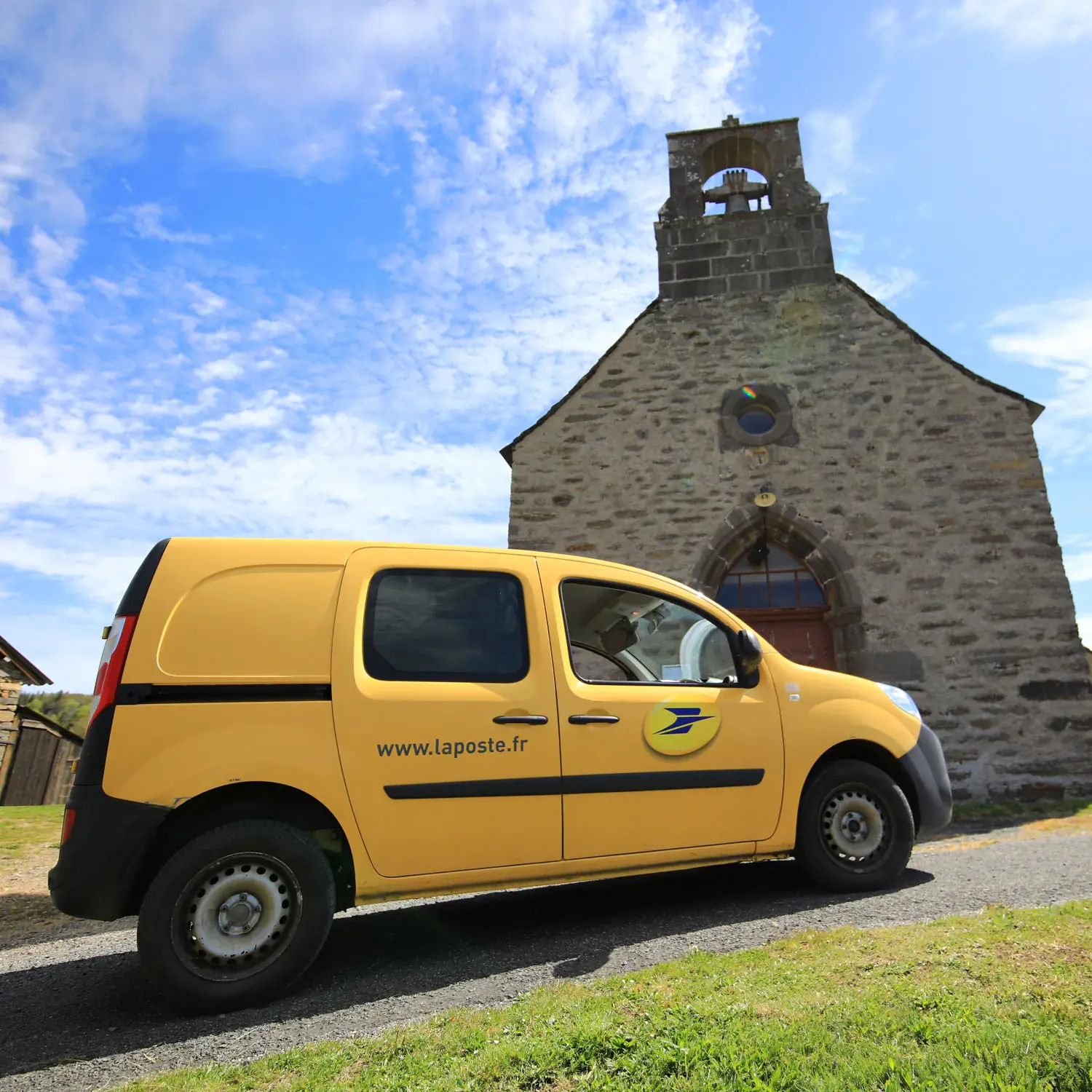 Yellow car flocked with the logo of La Poste parked in front of a chapel.