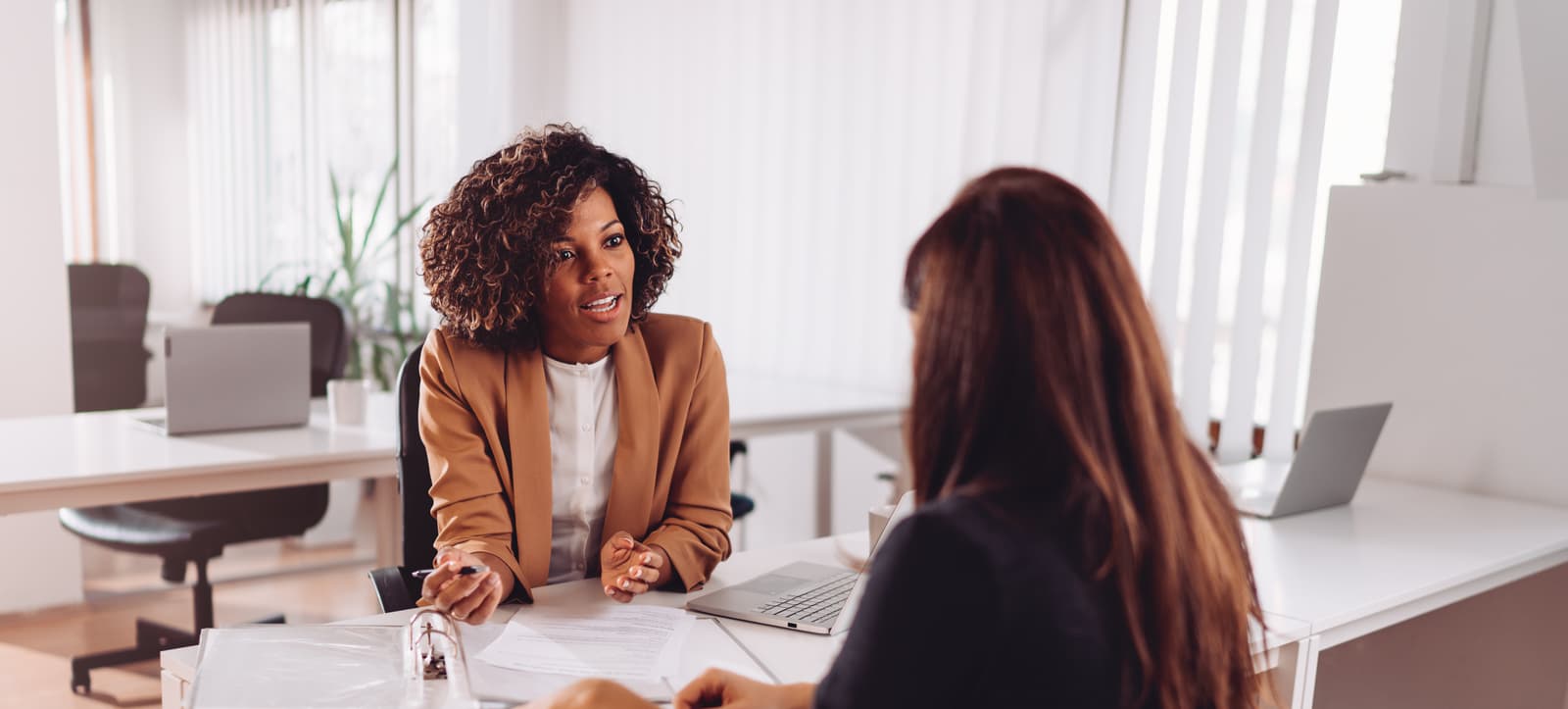 Two women talking around a desk, sitting in front of papers and a computer.