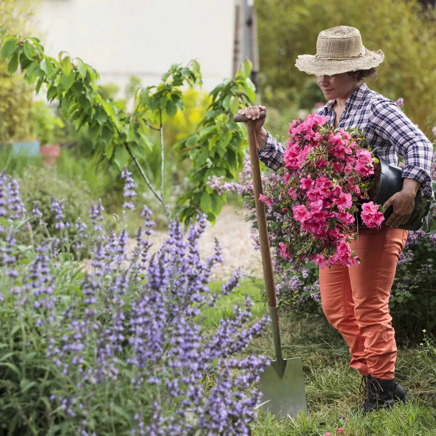 Woman wearing a hat and carrying a pot of flowers in her arm, gardening in an outdoor garden and holding a spade in her other hand.