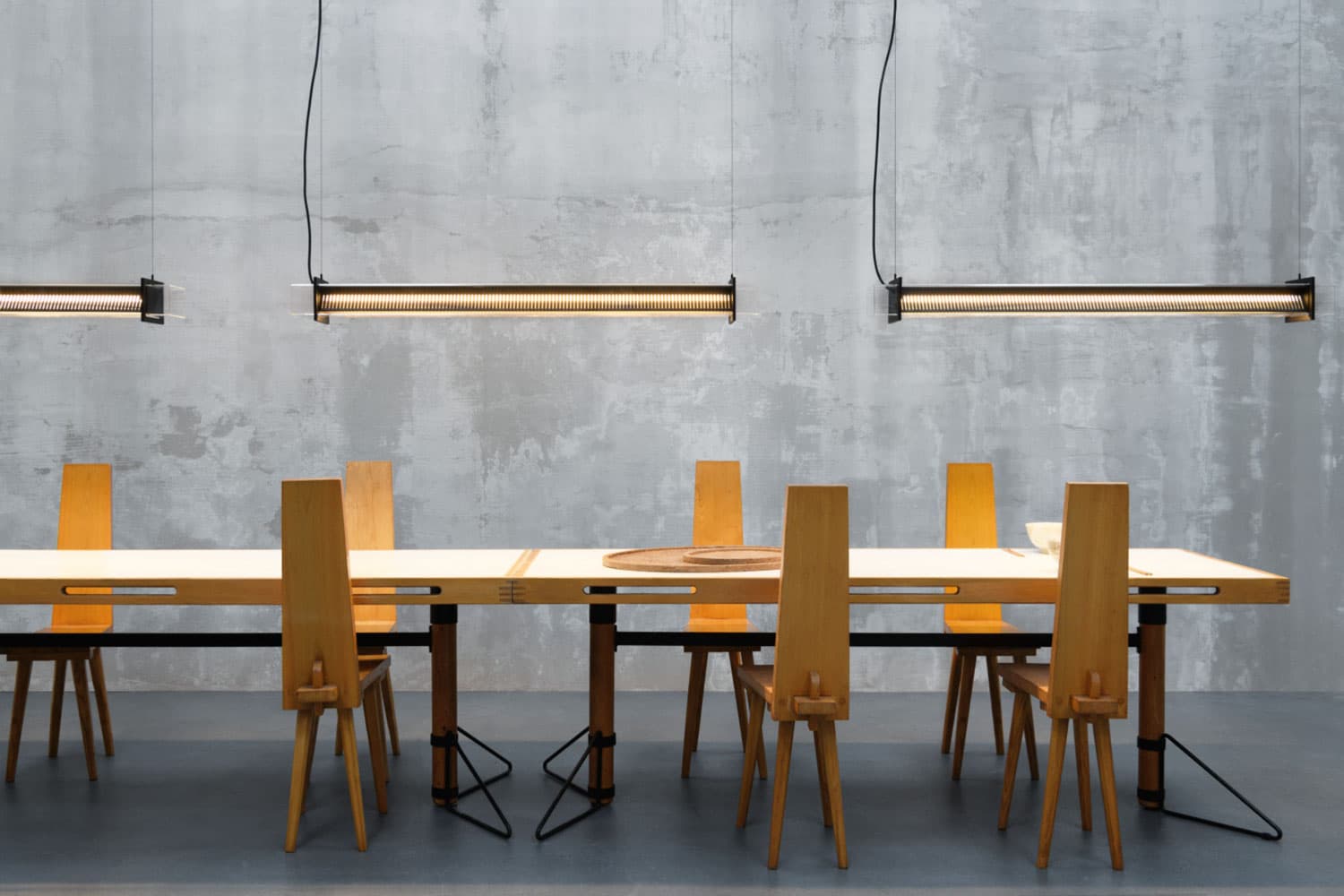 Large table in a concrete room with 7 chairs around it and a pendant light illuminating the entire table, by the brand Sammode