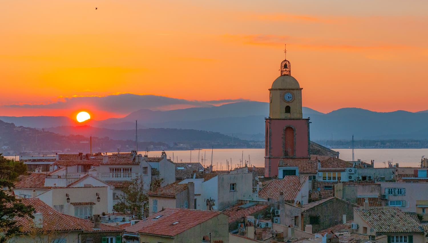 A blazing sunset over the Provençal rooftops of Saint-Tropez, highlighting its famous pink and yellow bell tower set against the sea and mountains.